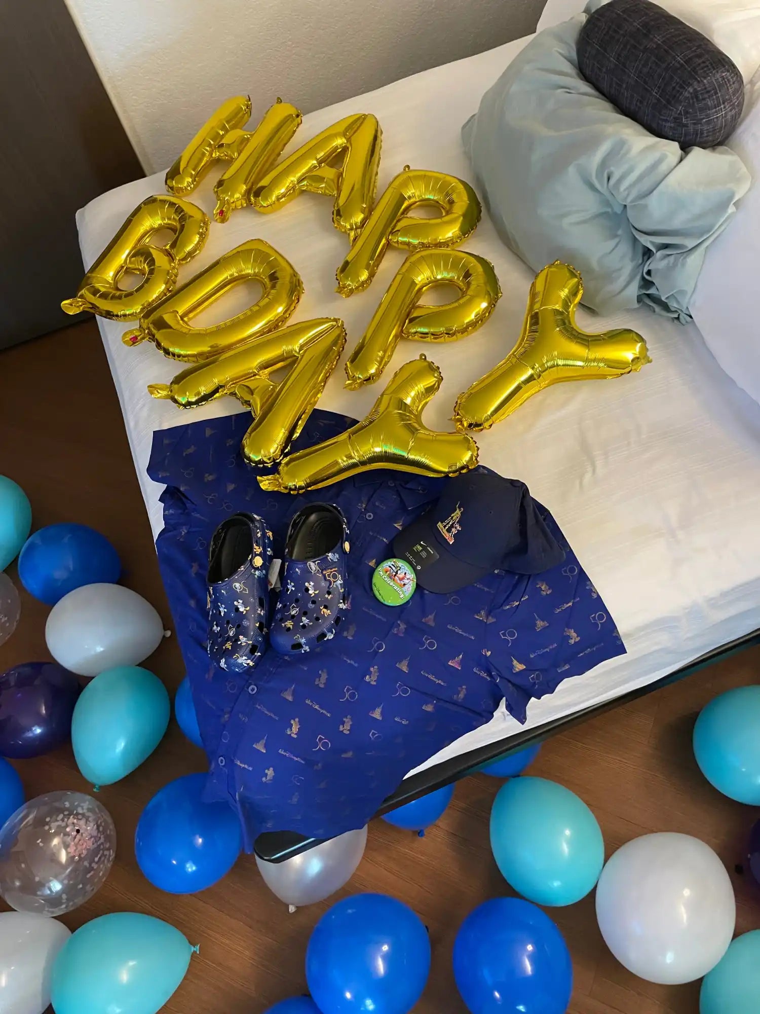 A collection of gold foil ’Happy Birthday’ balloons, a navy blue patterned shirt, a matching baseball cap, and a pair of navy blue Crocs.